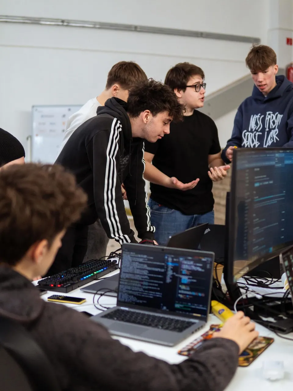 Hackathon HalovaMake: študenti pri notebookoch a monitoroch, Dušan stojí v strede v čiernom tričku a okuliaroch